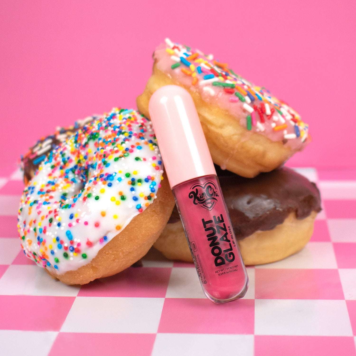 A playful composition featuring three donuts topped with colorful sprinkles and a pink lip gloss tube labeled 'Donut Glaze' on a checkered background.