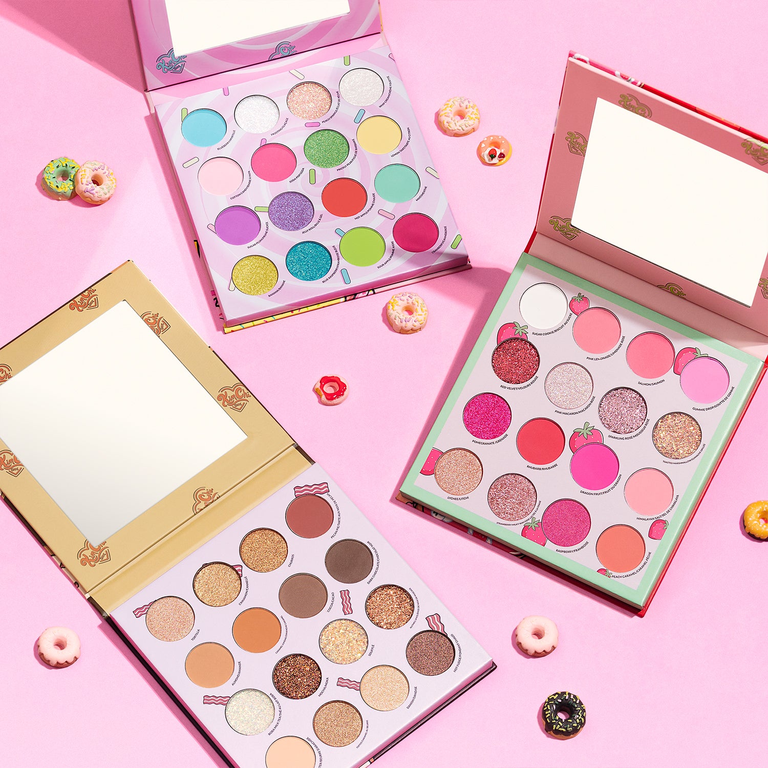 Three colorful eyeshadow palettes on a pink background, surrounded by small decorative donuts.