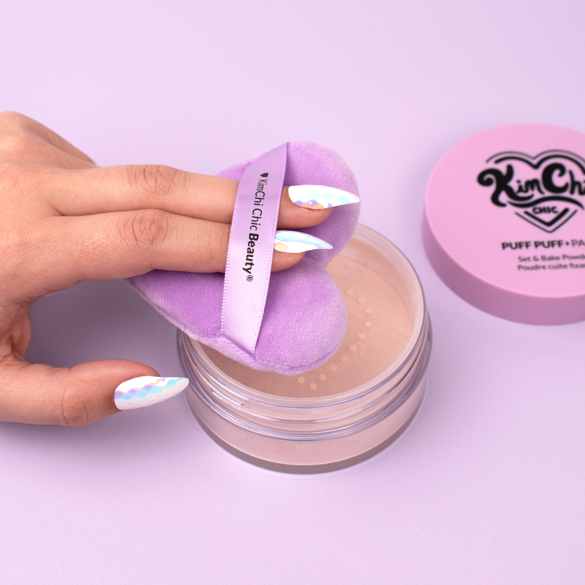 Hand using KimChi Chic Beauty Puff Puff Pass setting powder with a lavender applicator, in stylish packaging.