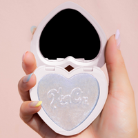 A hand holding a heart-shaped compact mirror with a shimmering light blue highlighter inside.