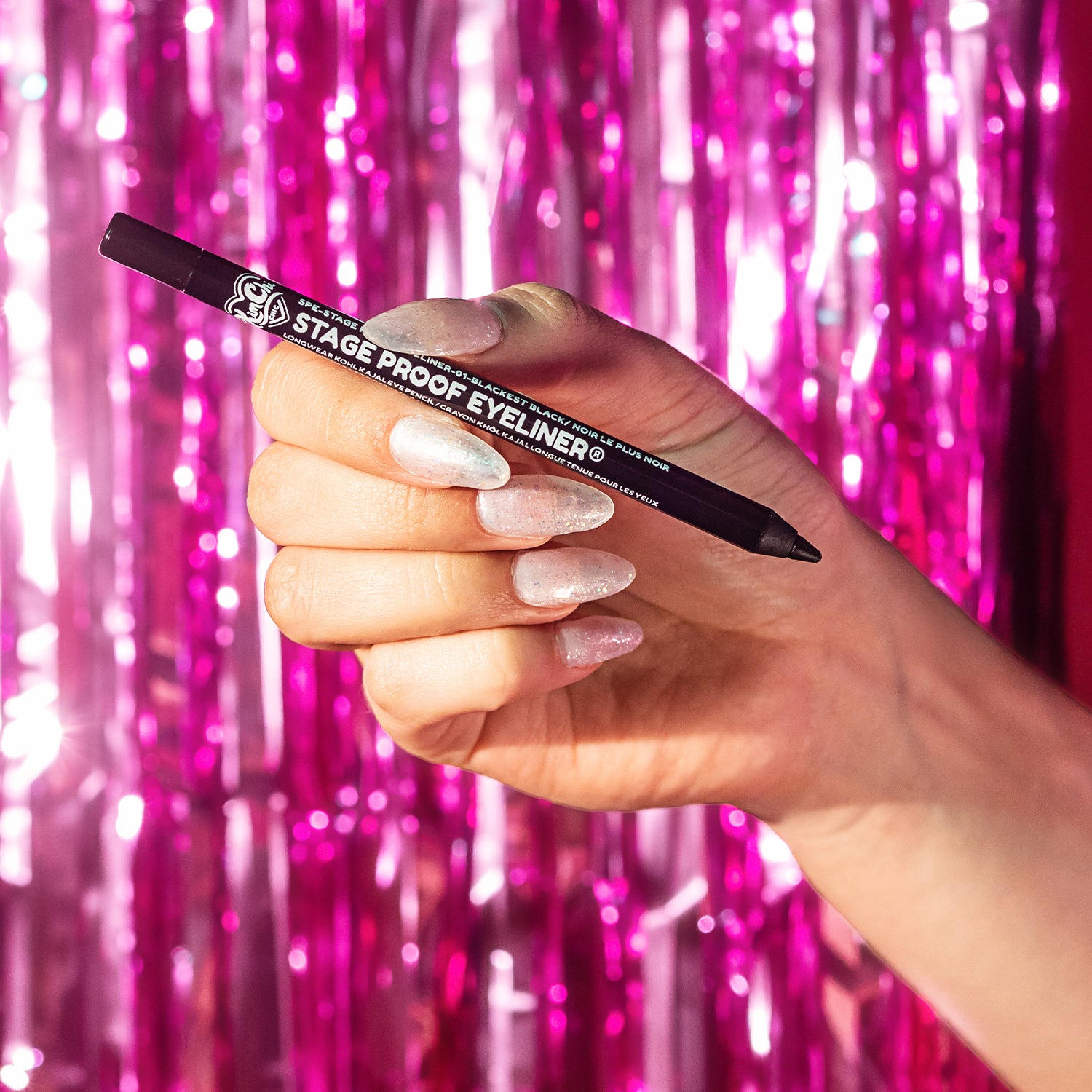 A hand holding a black eyeliner pencil against a shiny pink backdrop.