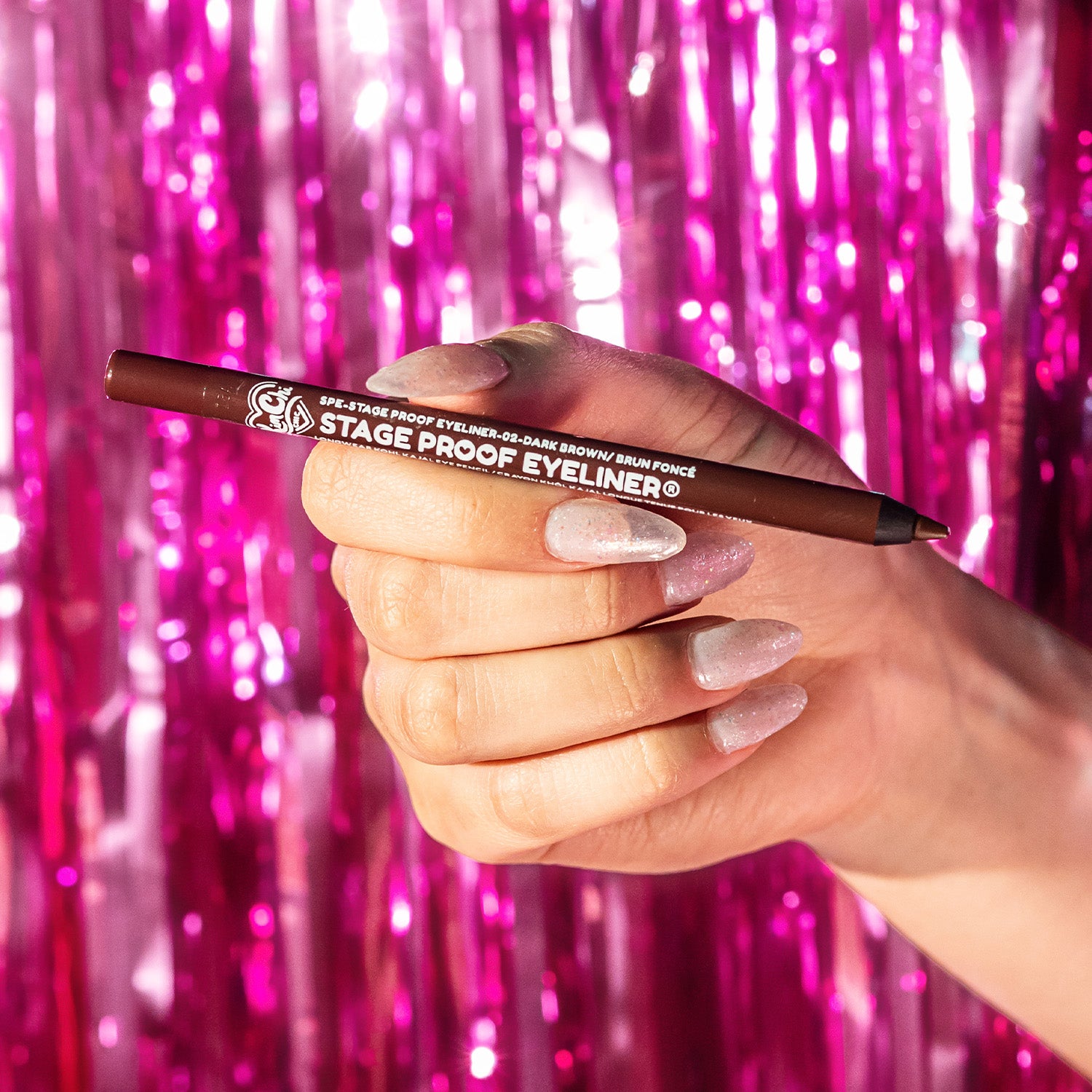 Person holding a brown eyeliner pencil in front of a sparkling pink backdrop.