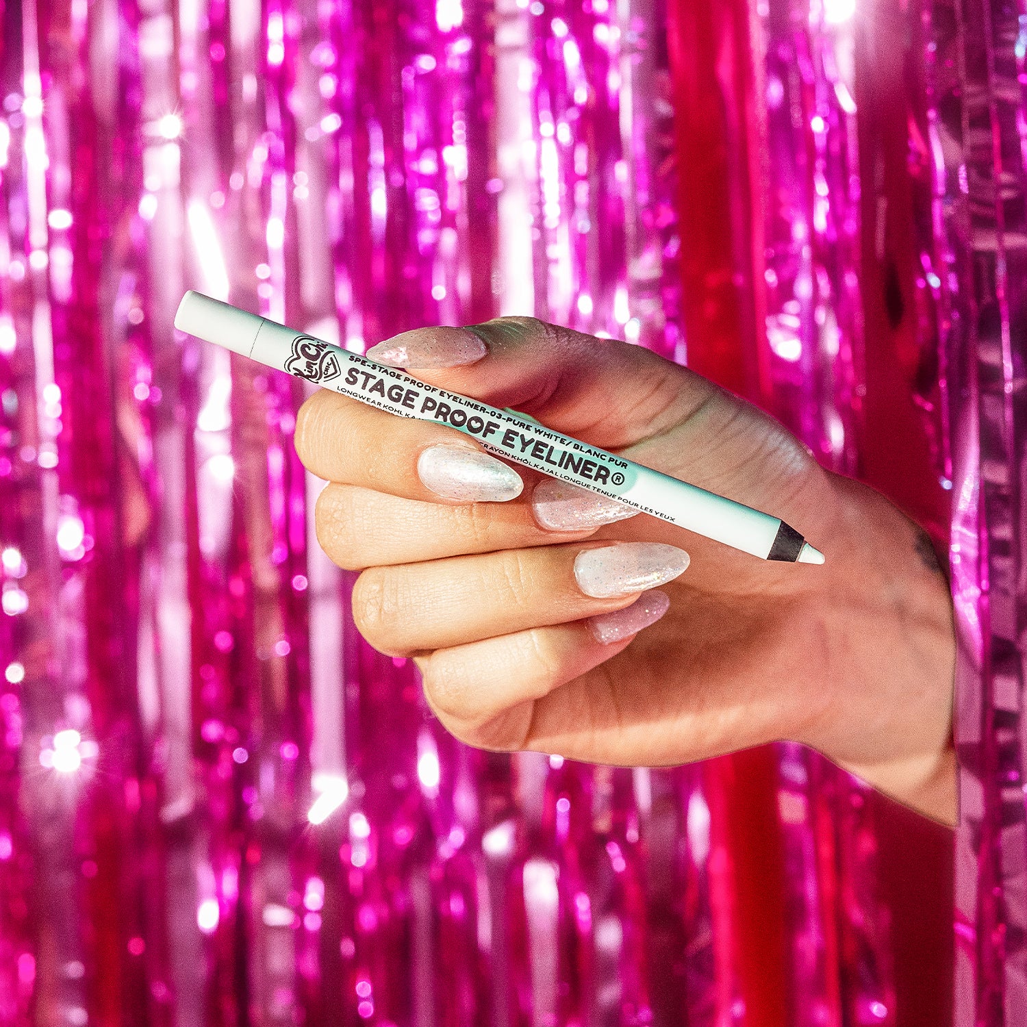 A hand holding a white eyeliner pencil in front of shiny pink tinsel curtains.
