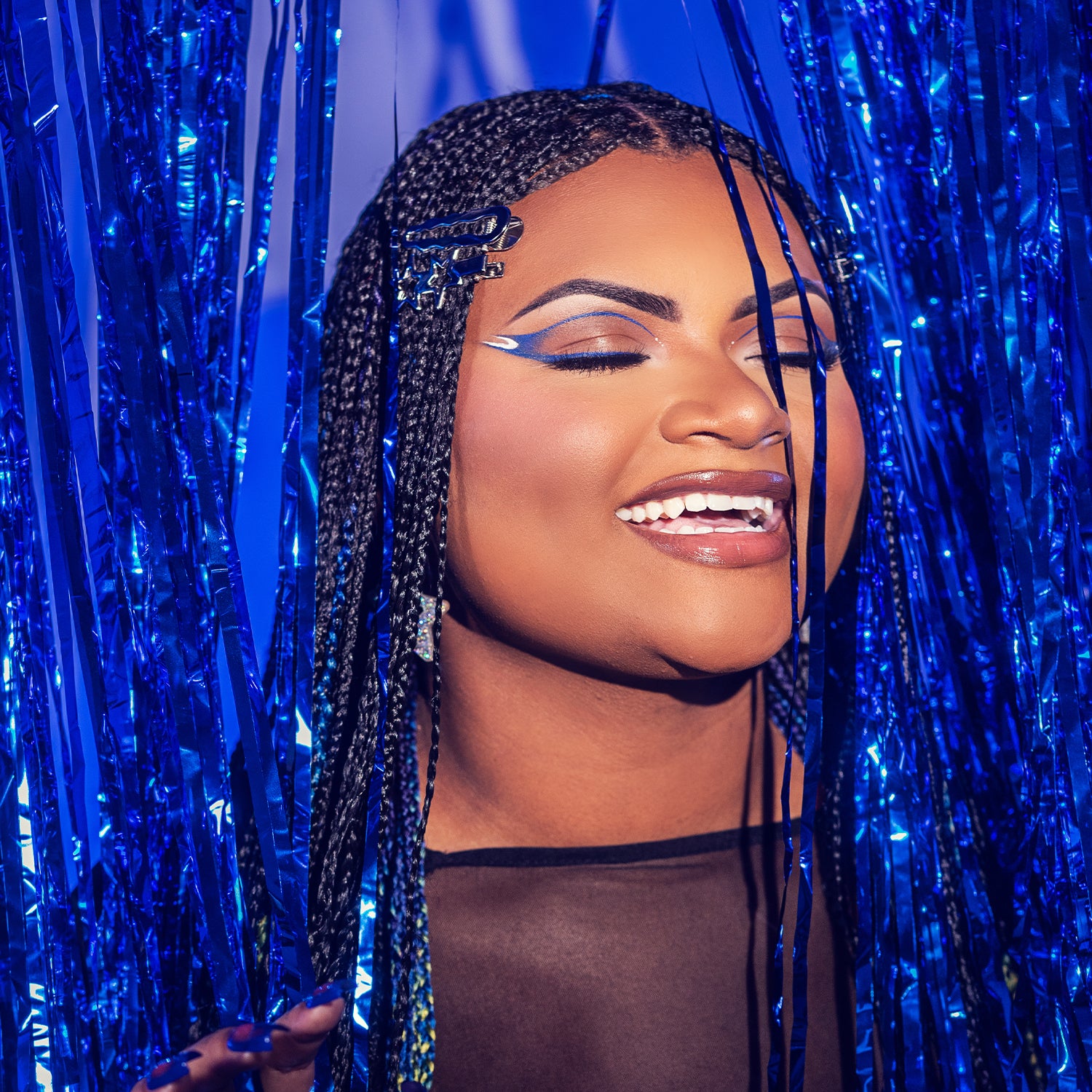 A person with braids smiling in front of blue metallic fringe.