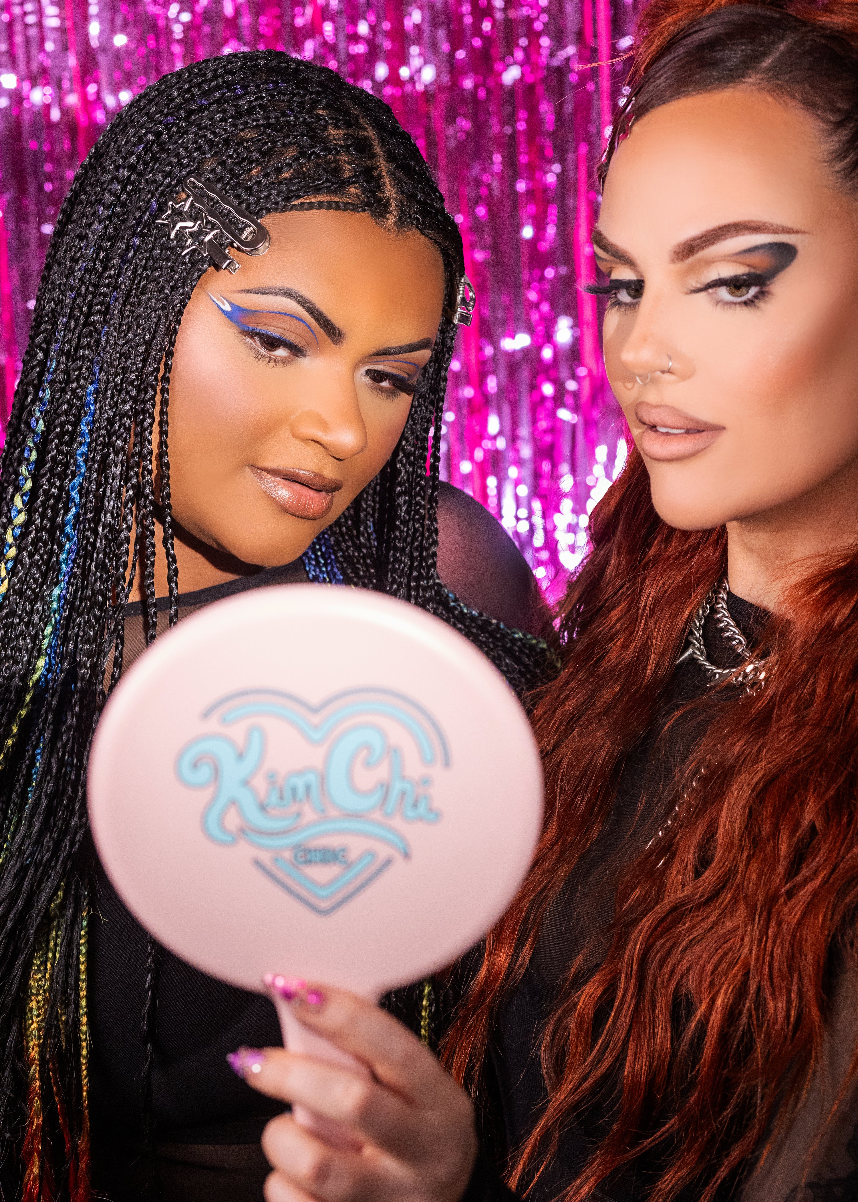 Two individuals posing with a beauty mirror featuring the brand logo, set against a colorful, sparkly background.