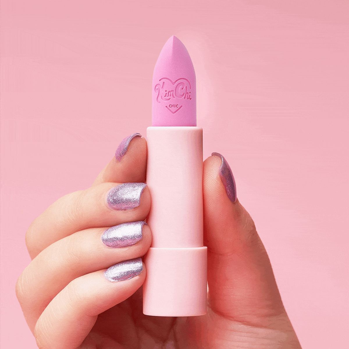 A hand holding a pink lipstick against a pastel pink background.