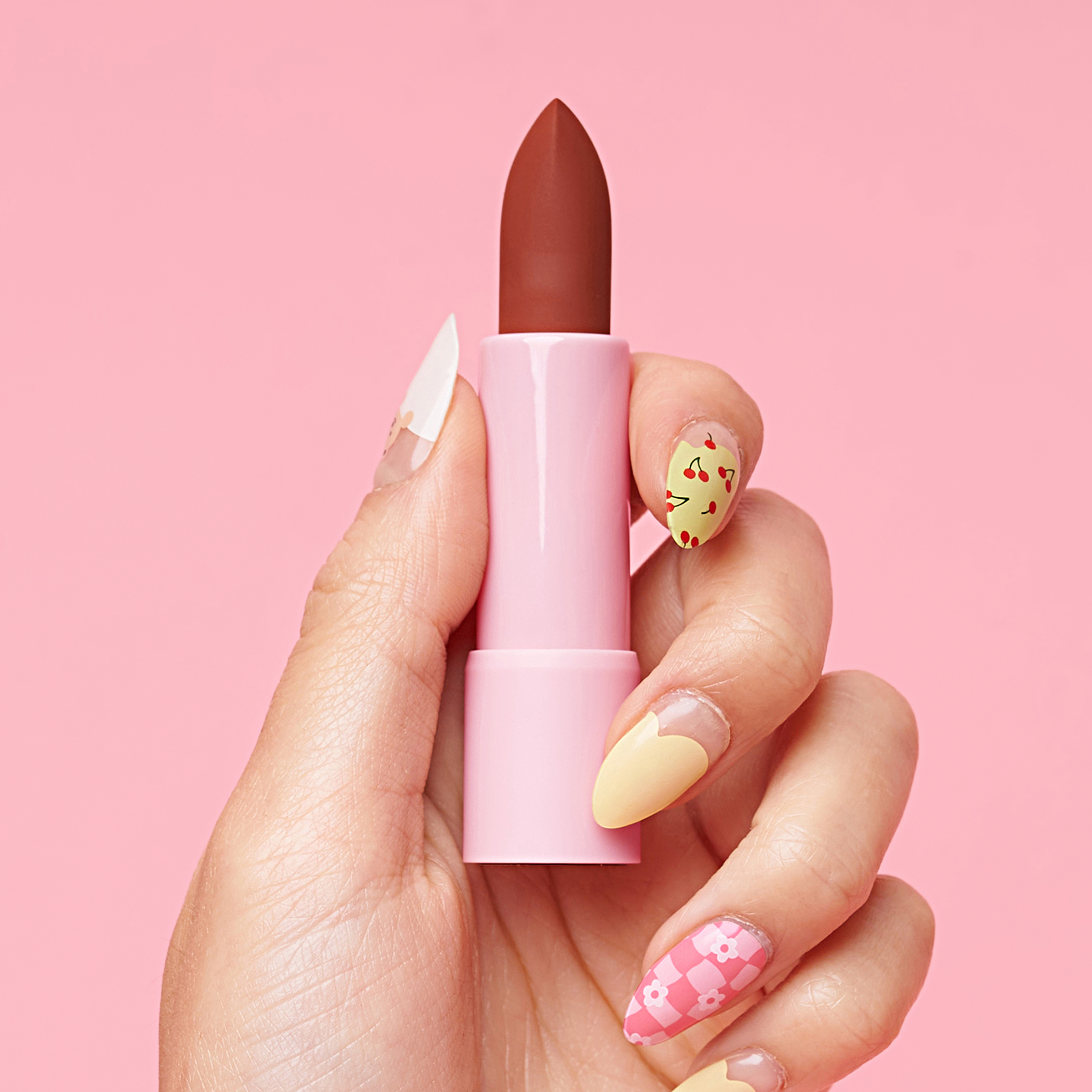 Hand with nail art holding a pink lipstick against a pink background.