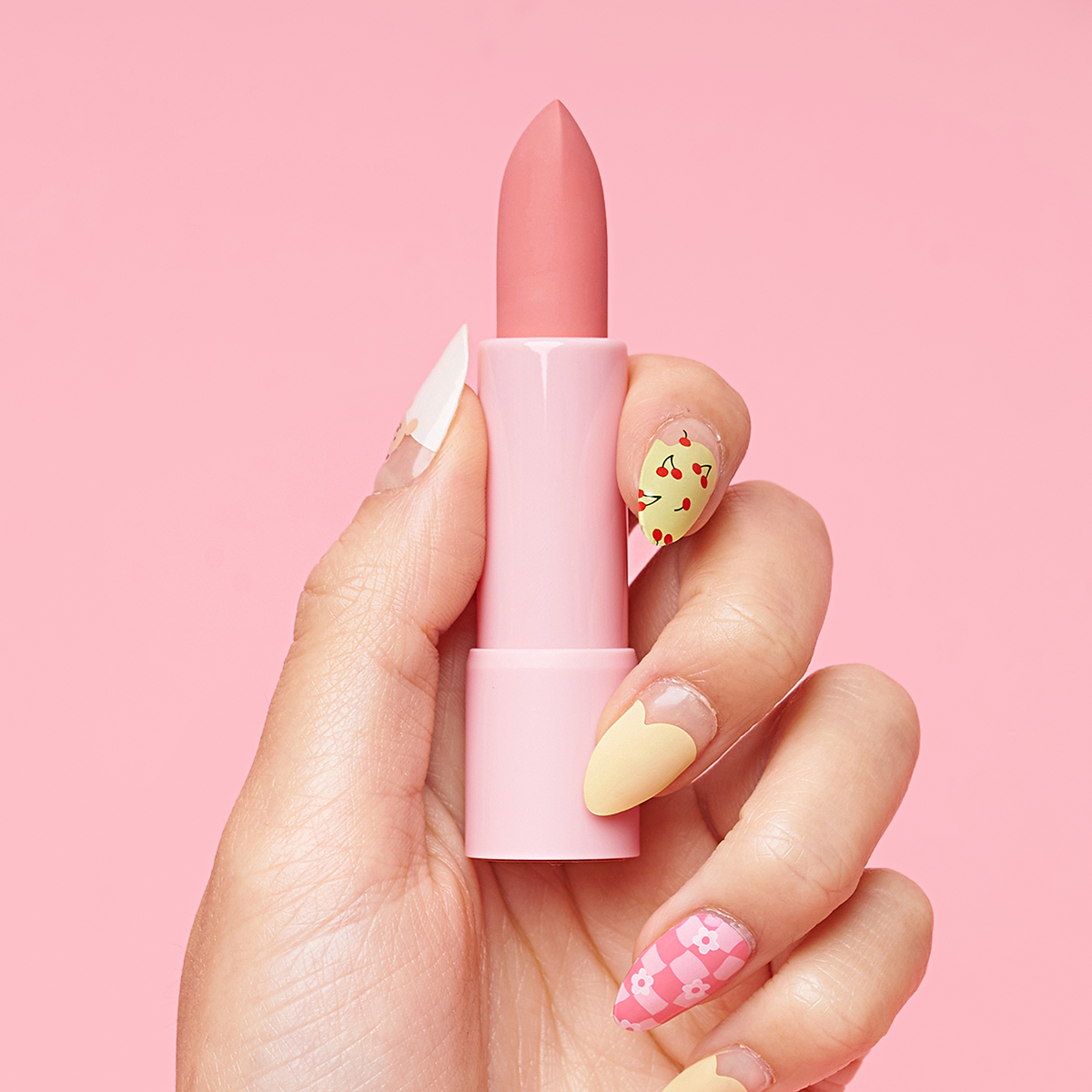 Hand with colorful nails holding pink lipstick against a pink background