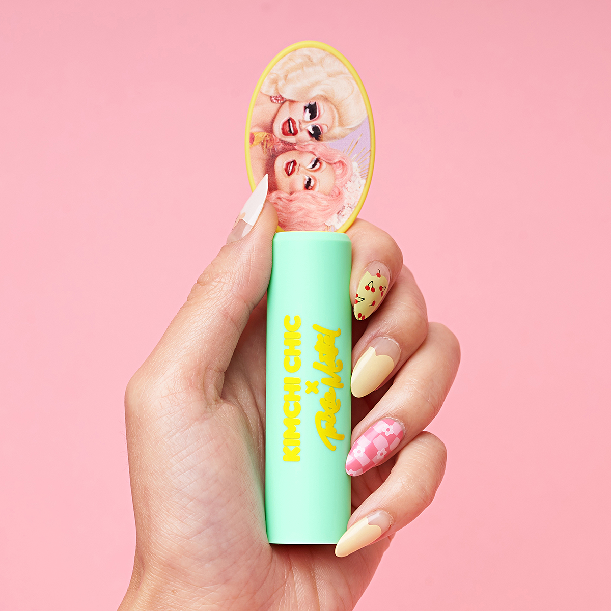 Hand holding a mint-green lipstick with decorative top against a pink background.