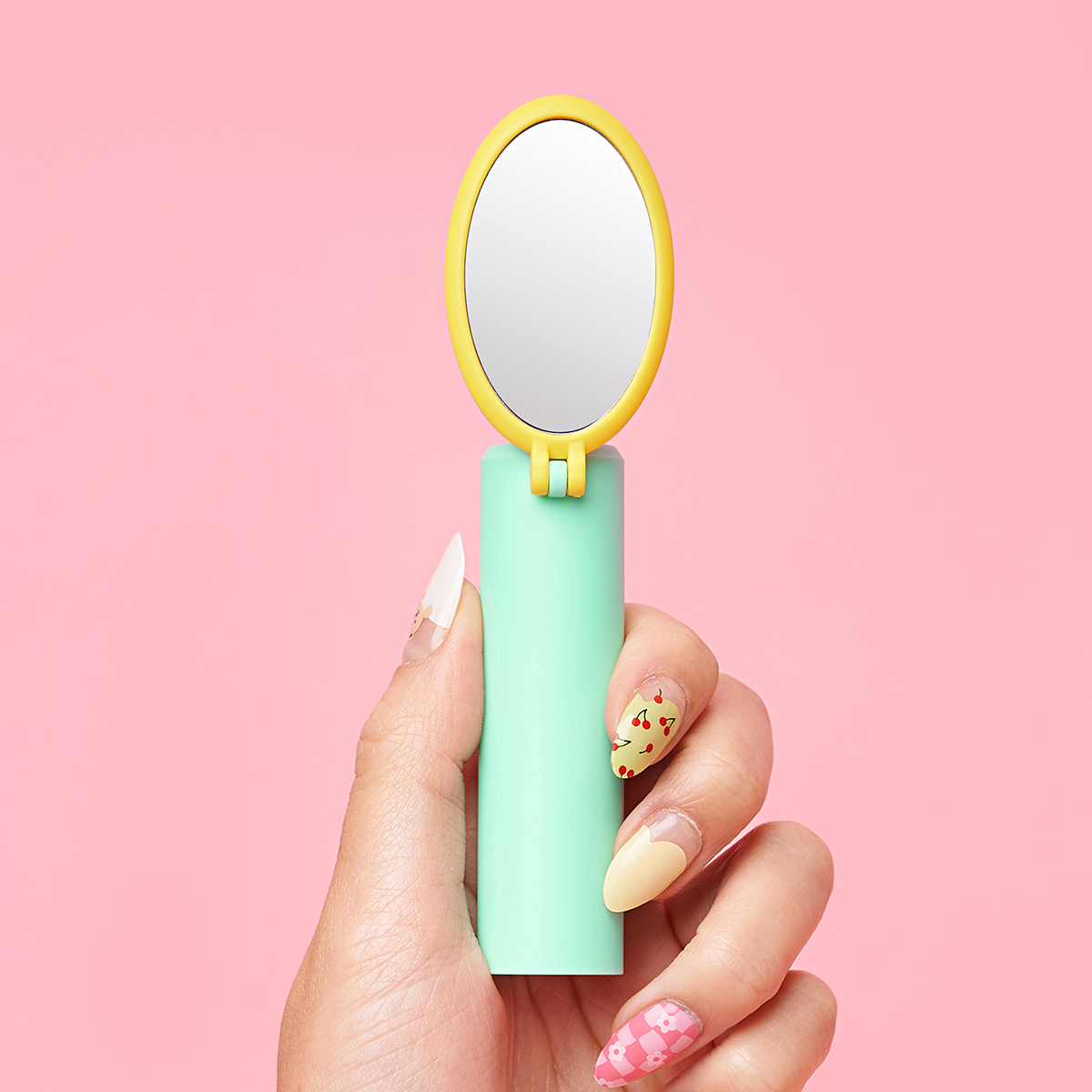 Hand holding a compact mirror with pastel nails against a pink background.