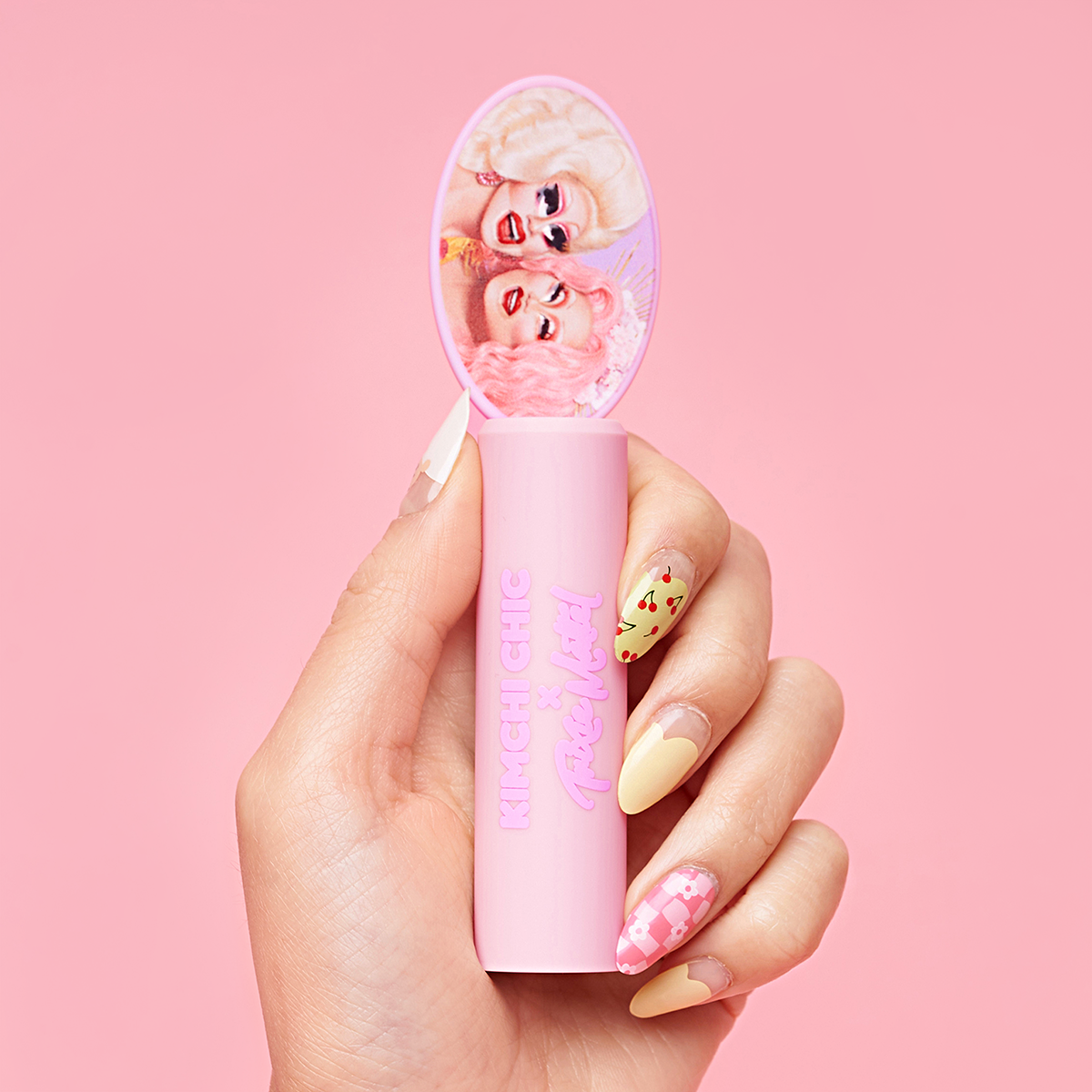 Hand holding a pink cosmetic product with illustrated figures on the cap against a pink background.