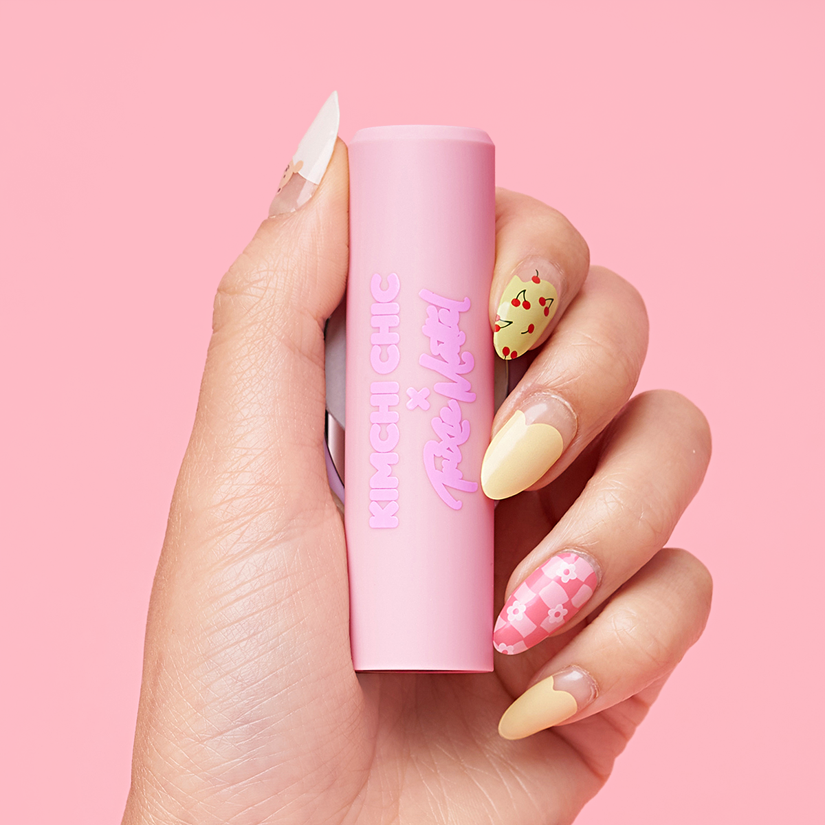 Hand holding a pink KimChi Chic cosmetic tube with decorative nails on a pink background.