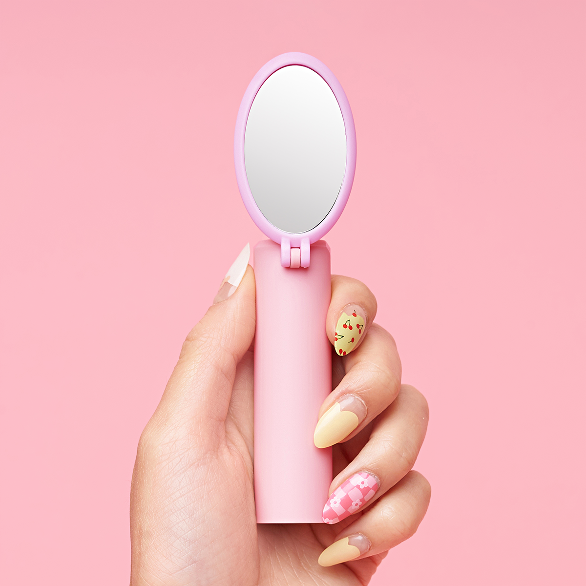 Hand holding a pink compact mirror with decorated nails on a pink background