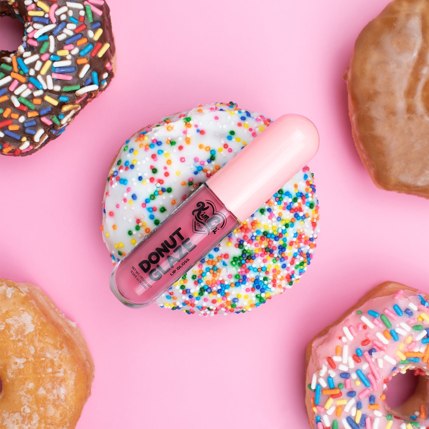 A lip gloss tube resting on a donut covered in white icing and colorful sprinkles, surrounded by other donuts on a pink background.