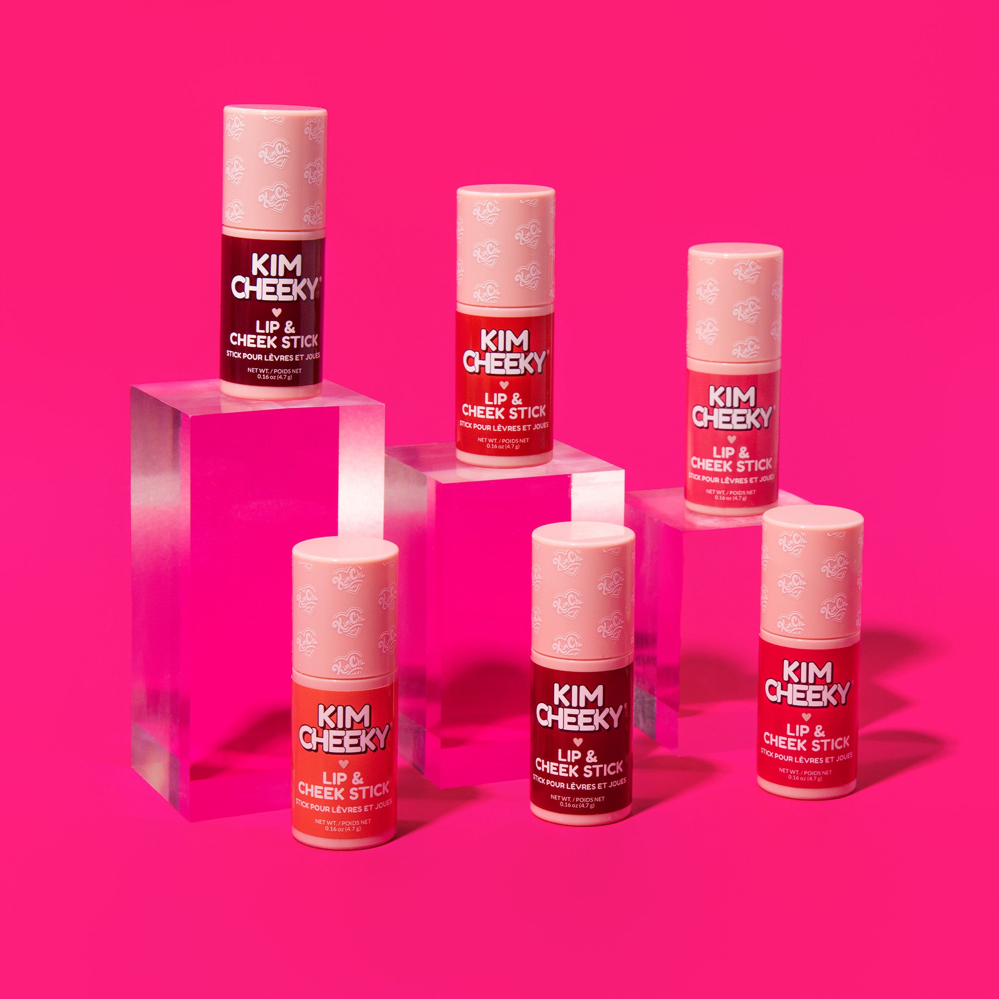 Five colorful lip and cheek sticks on clear acrylic pedestals against a bright pink background.