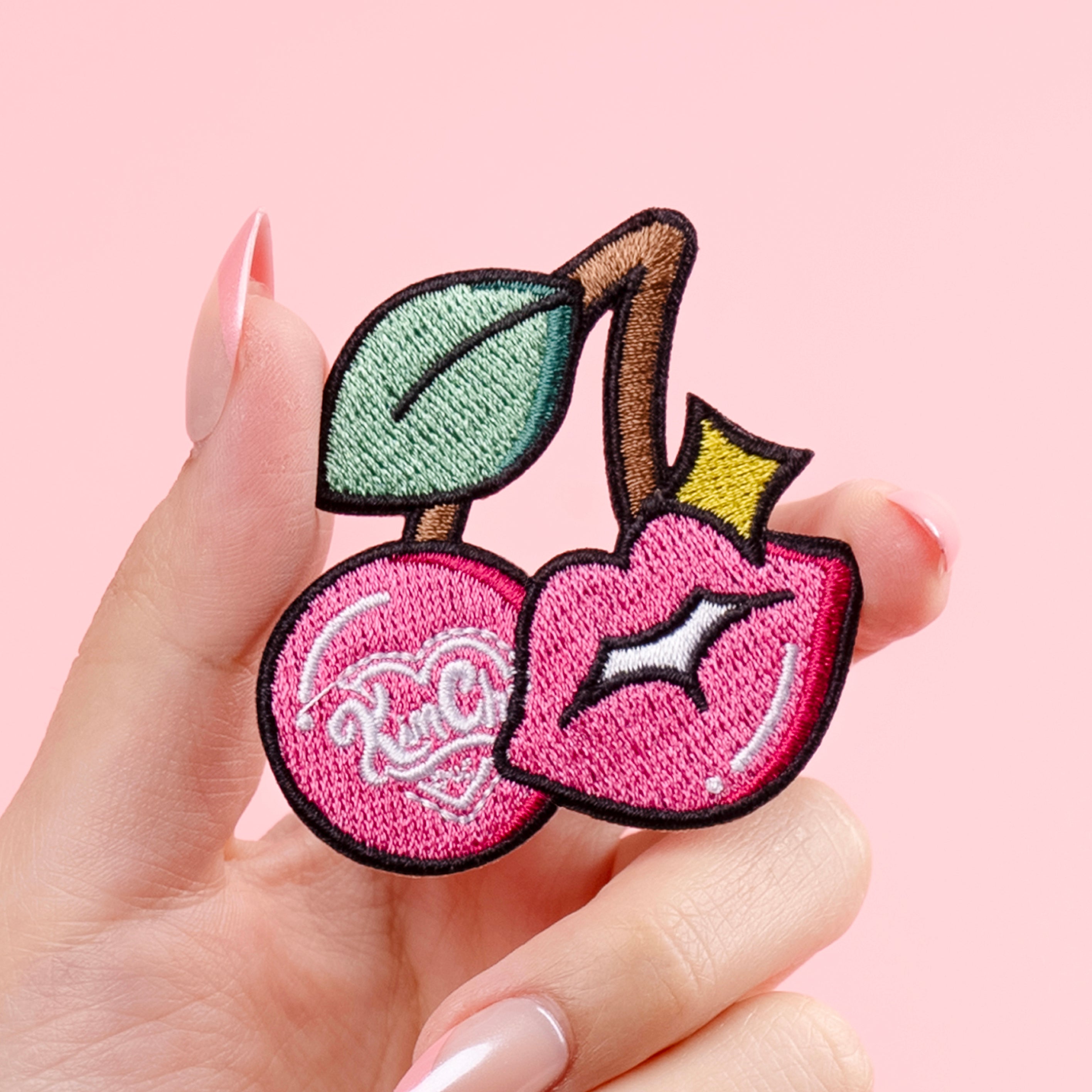 Person holding a pink cherry-shaped embroidered patch with a green leaf on a pink background.