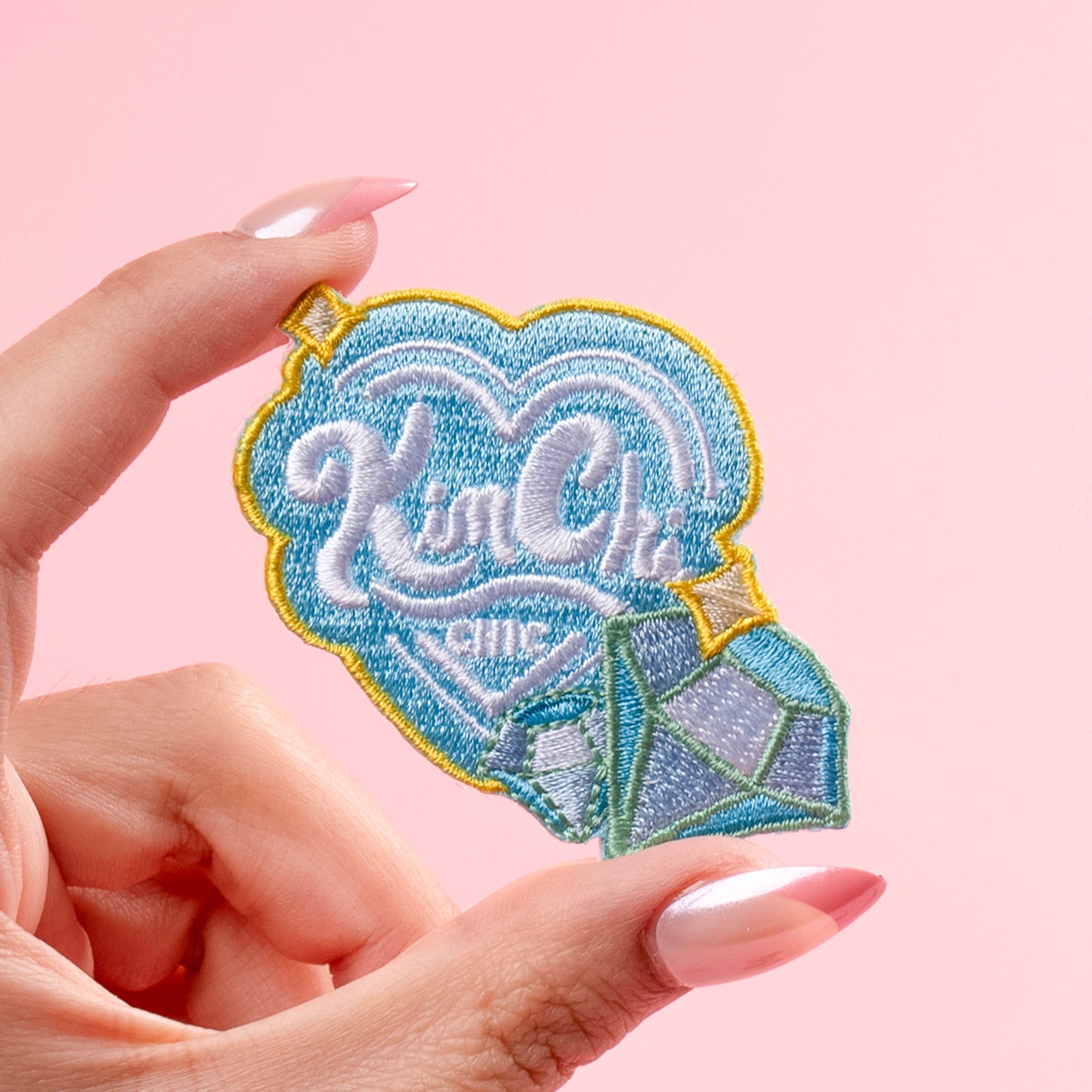 Hand holding a blue heart-shaped embroidered patch with decorative text and diamond design on a pink background.