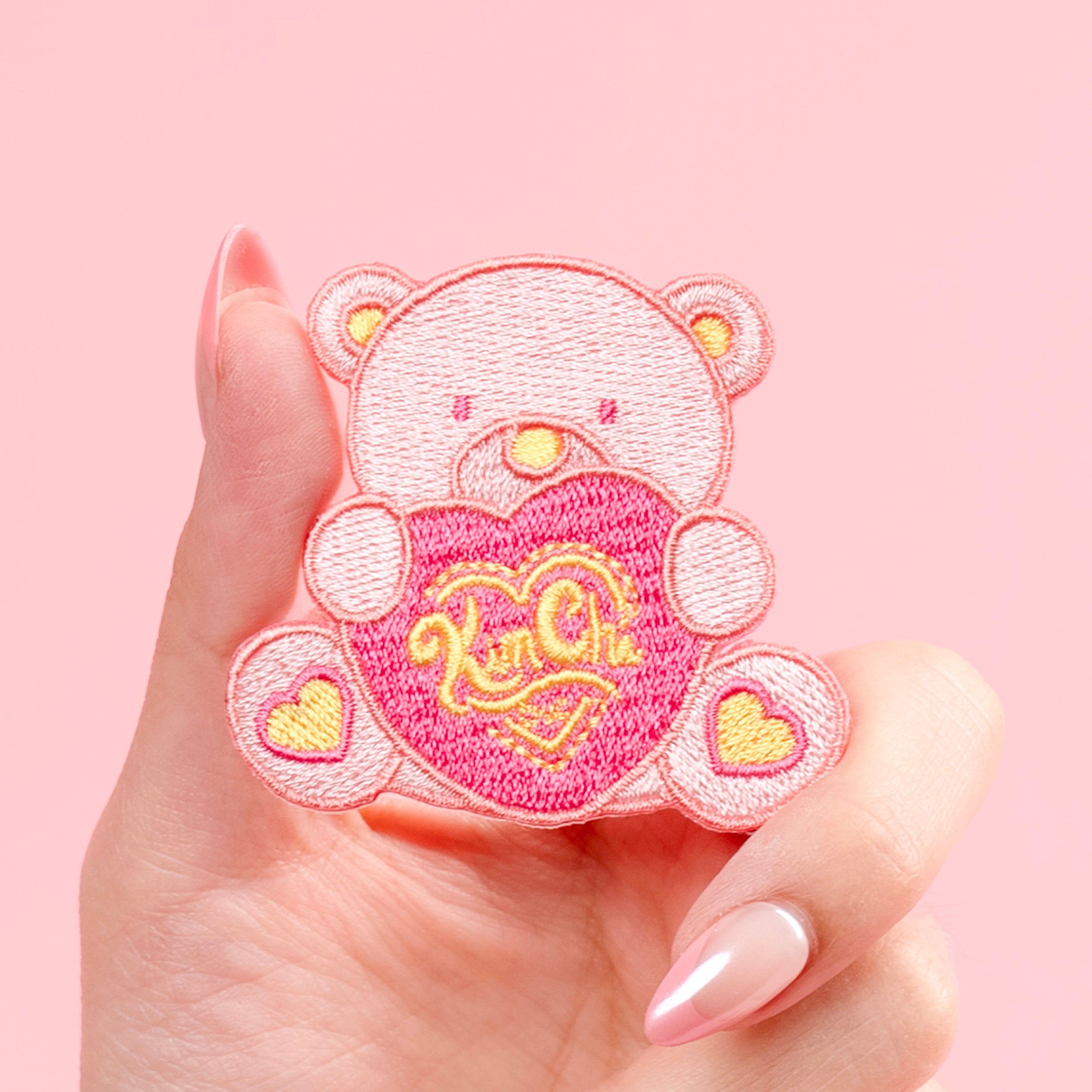 Hand holding a pink bear patch with a heart design.