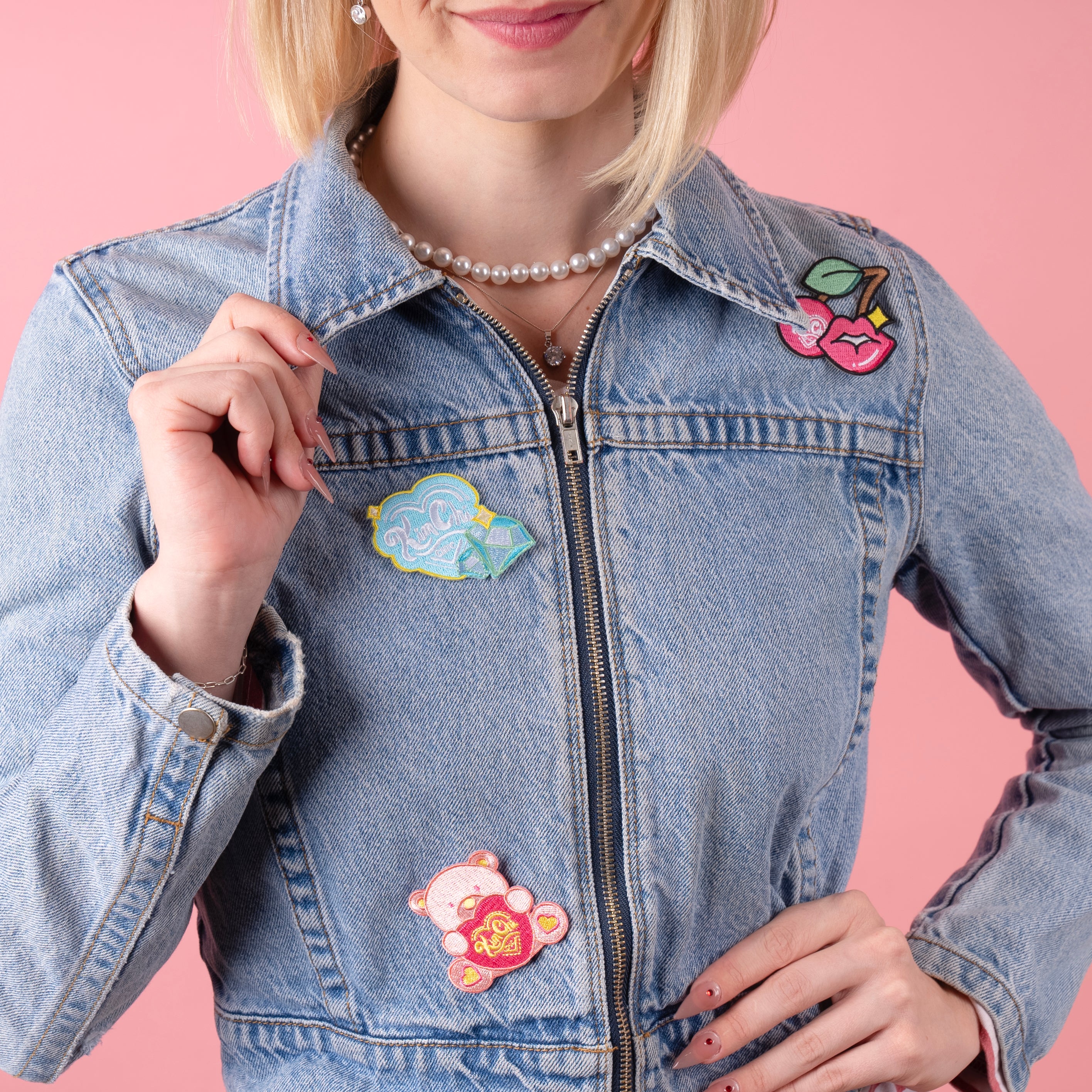 Person wearing a denim jacket with colorful embroidered patches on a pink background.