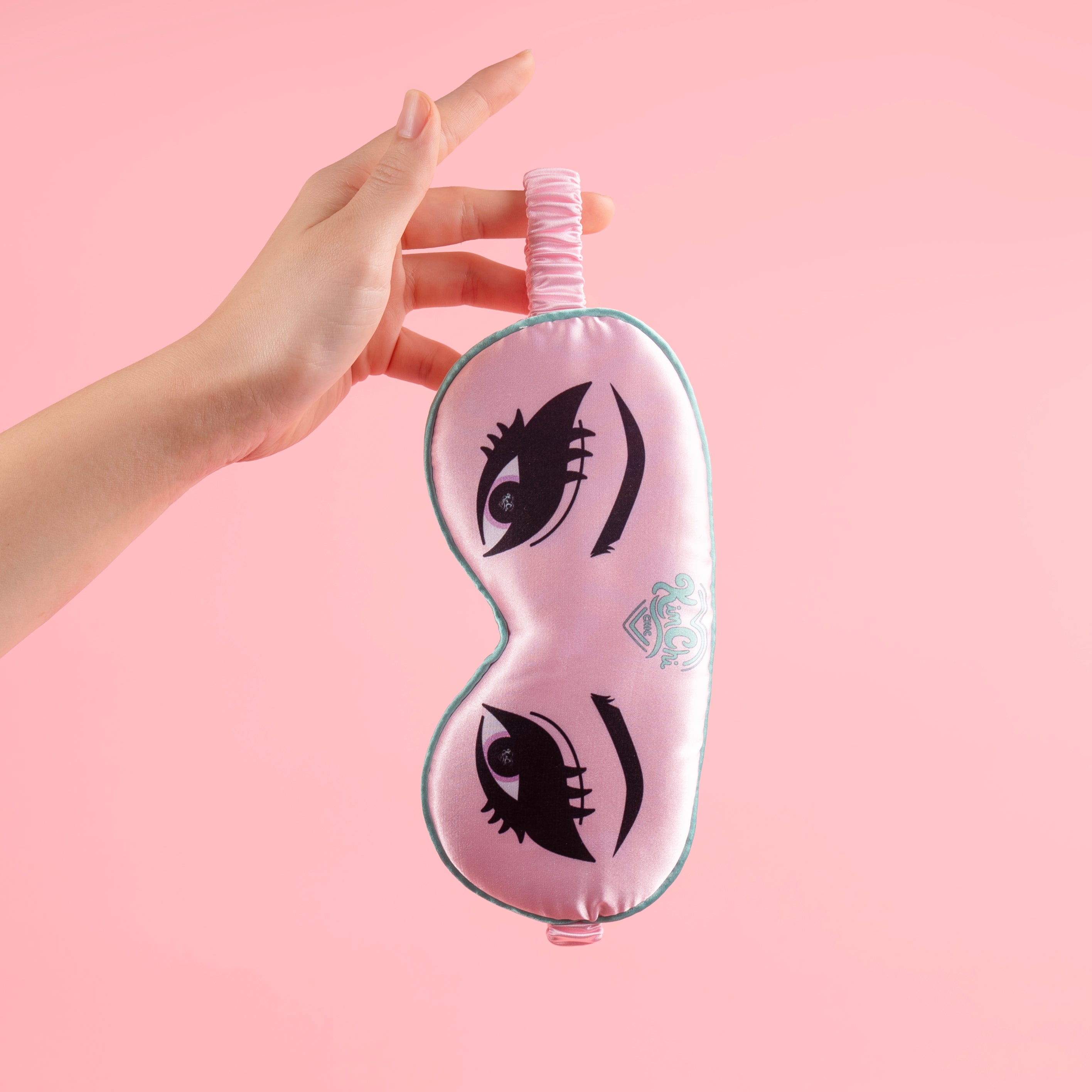 Hand holding pink sleep mask with eyelashes design on pink background