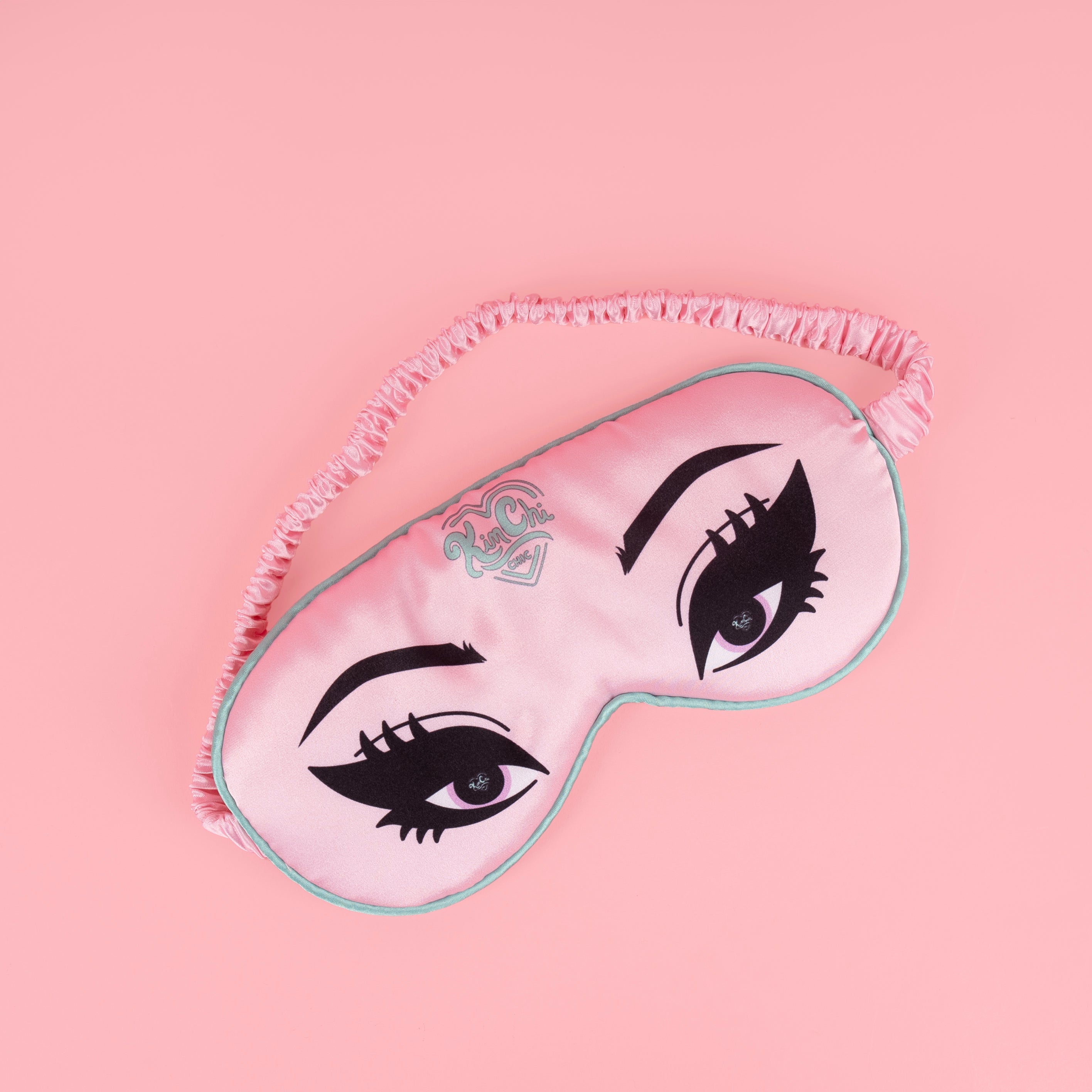 Pink sleep mask with stylized eyes design on a pink background.