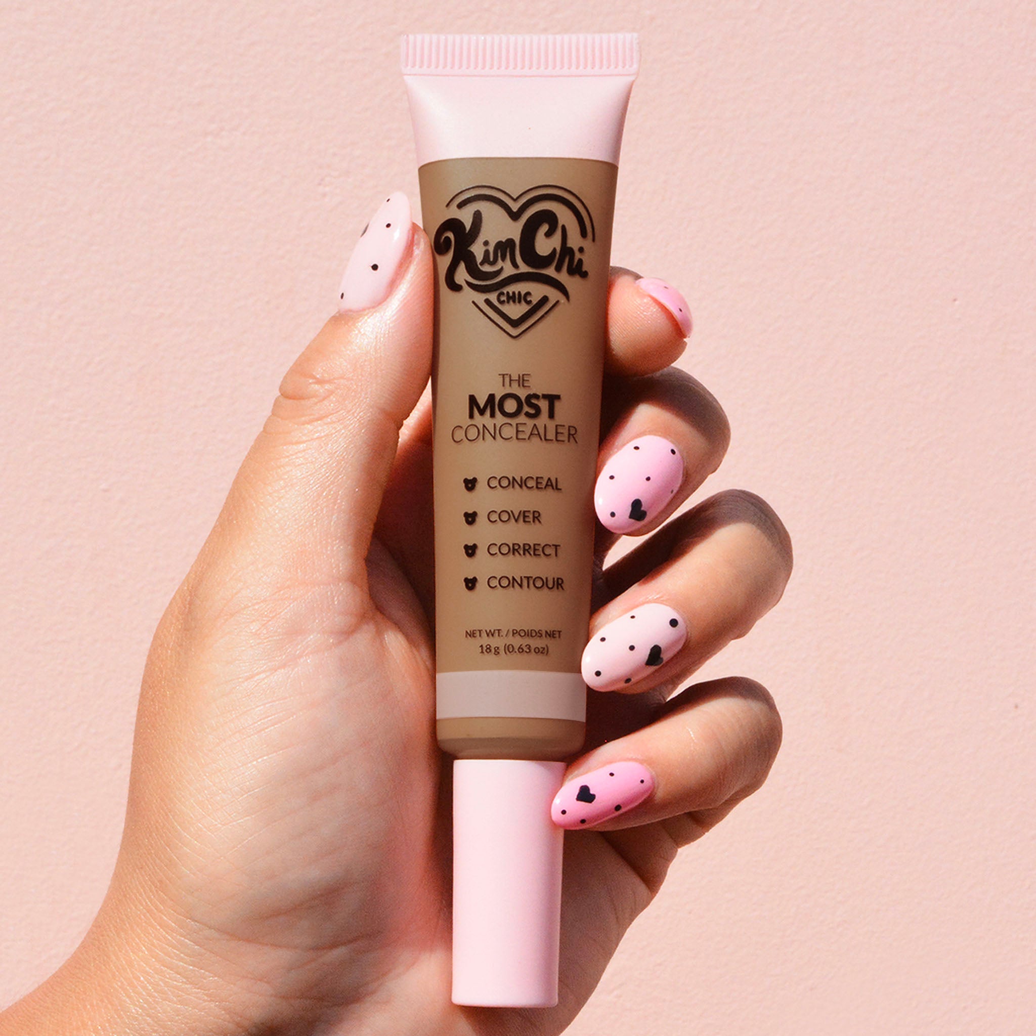 Hand holding a tube of Kim Chi Chic The Most Concealer against a pink background