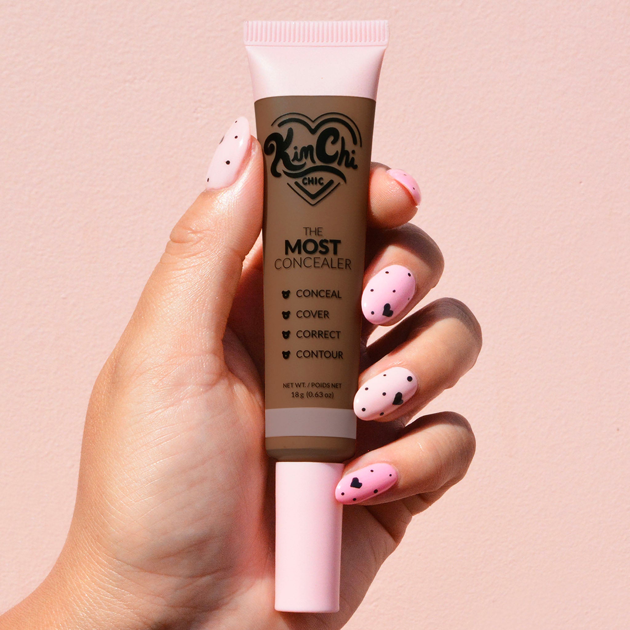 Hand holding a tube of Kim Chi The Most Concealer against a pink background.