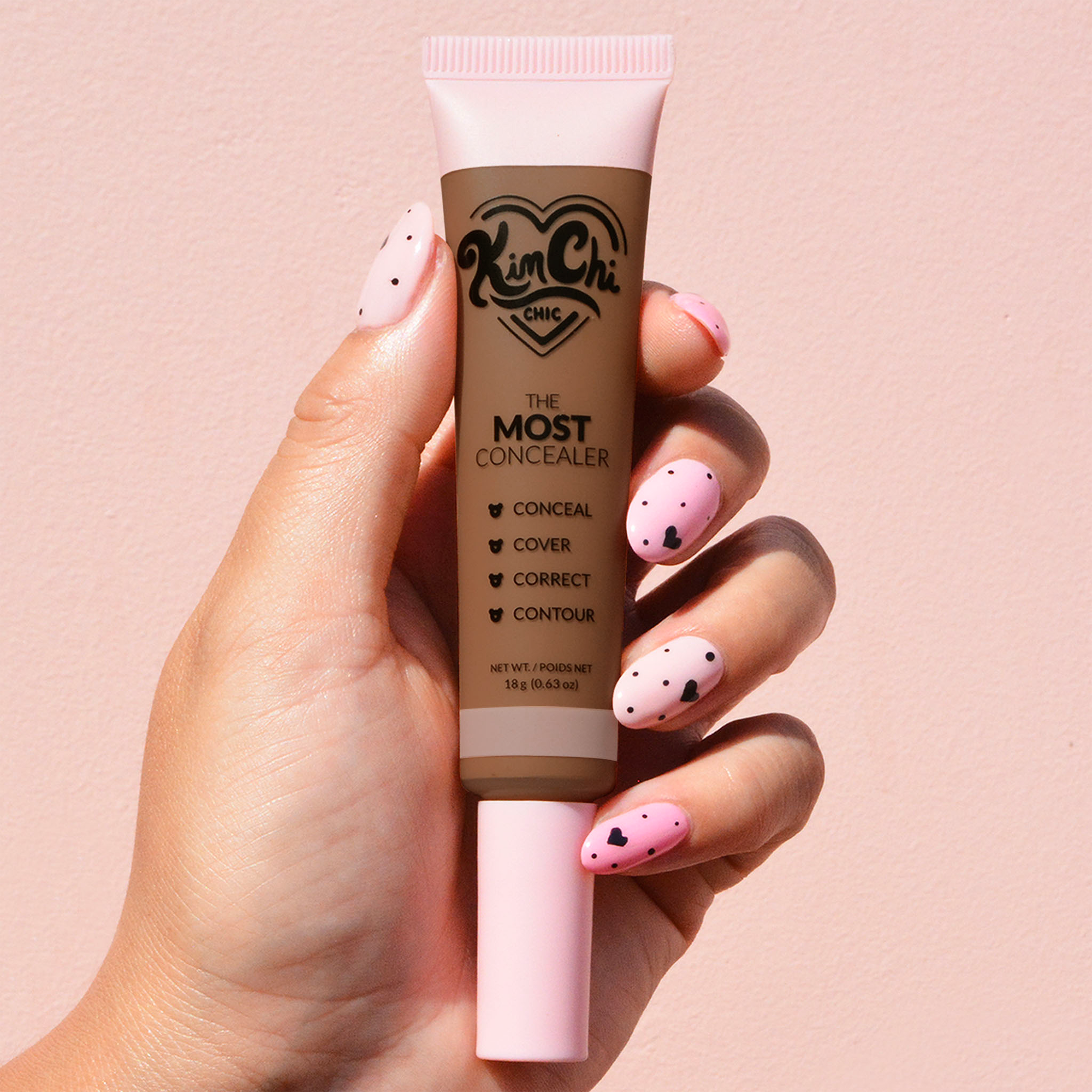 Hand holding a tube of concealer with pink nail polish against a pink background.