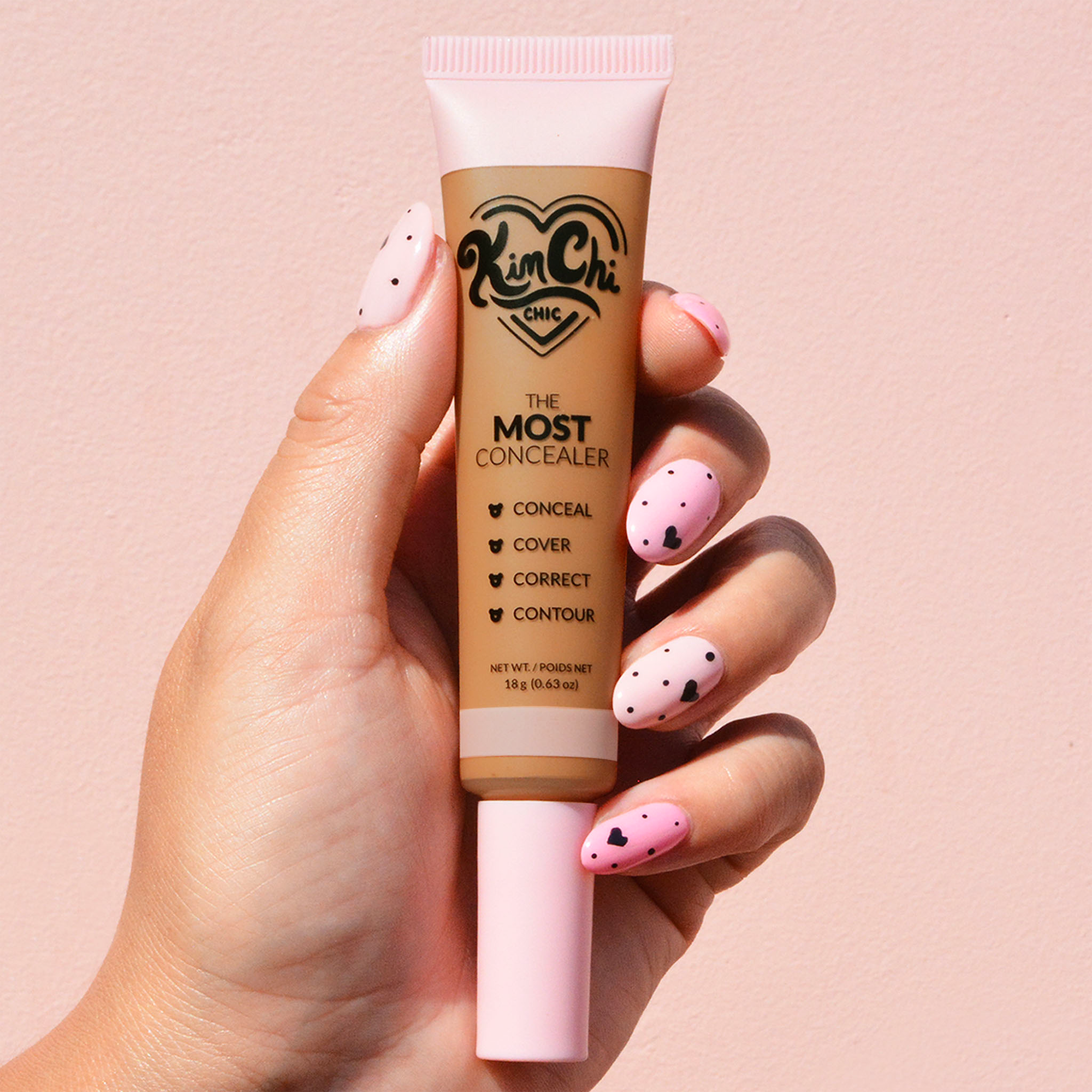 Hand holding a tube of Kim Chi Chic The Most Concealer against a pink background.