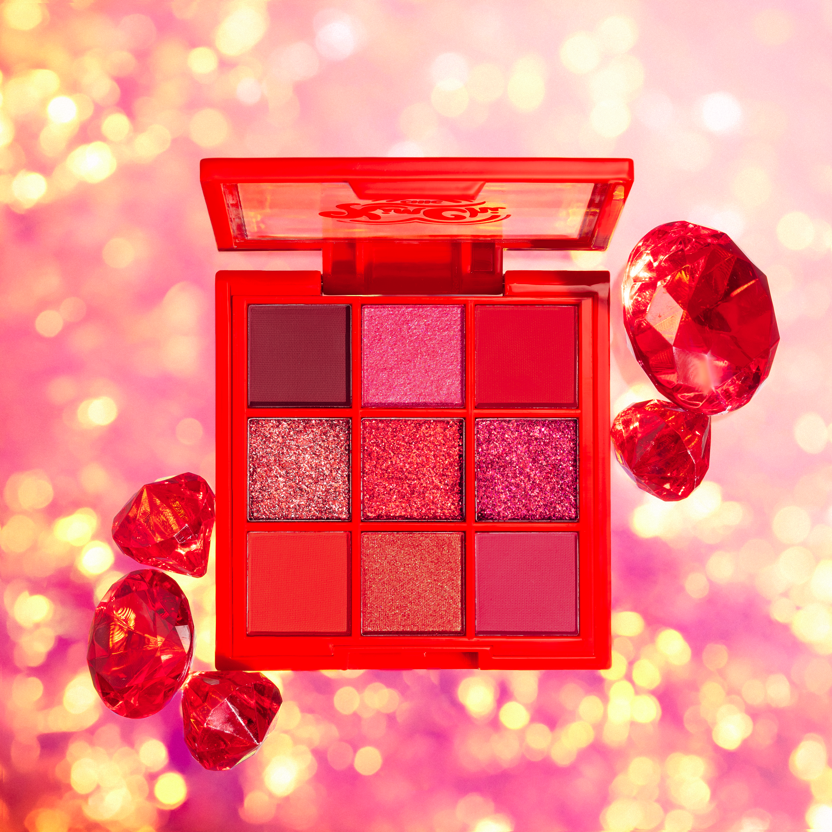 Red-themed eyeshadow palette surrounded by ruby gemstones on a pink sparkling background