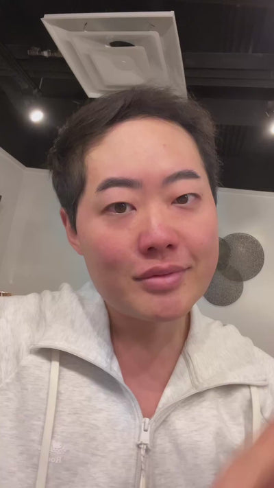 A person with short dark hair wearing a gray zip-up hoodie, sitting indoors and looking at the camera.