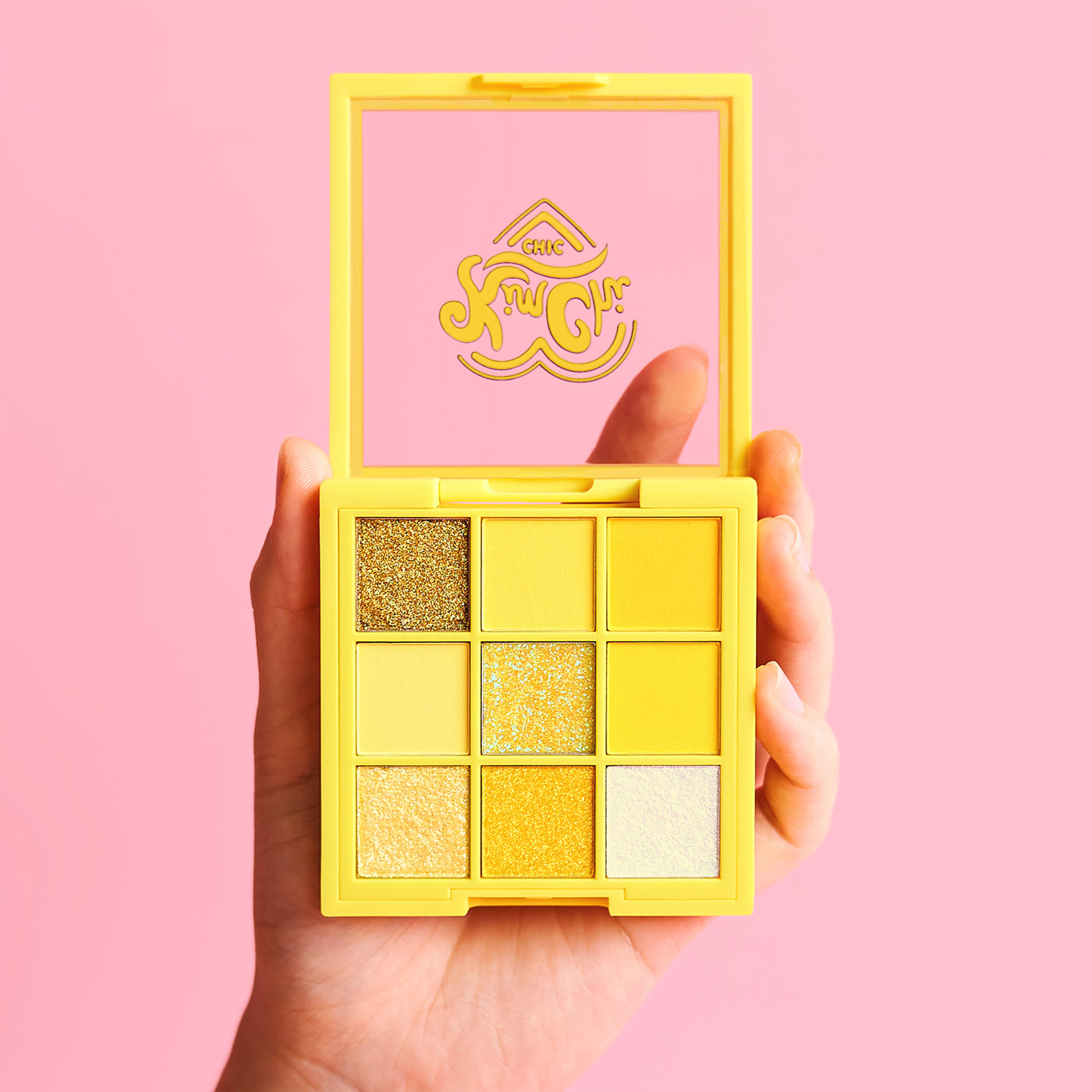 Hand holding a yellow eyeshadow palette with nine shades against a pink background.