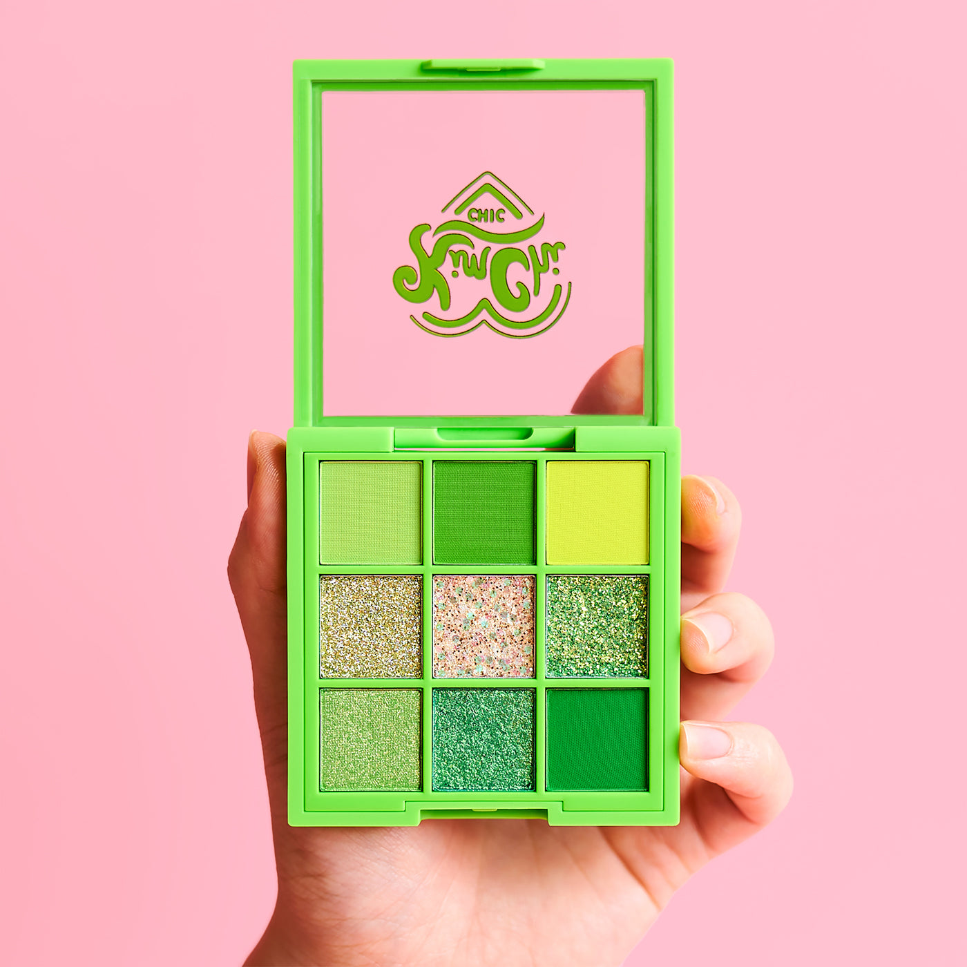 Hand holding a green eyeshadow palette with various shades against a pink background