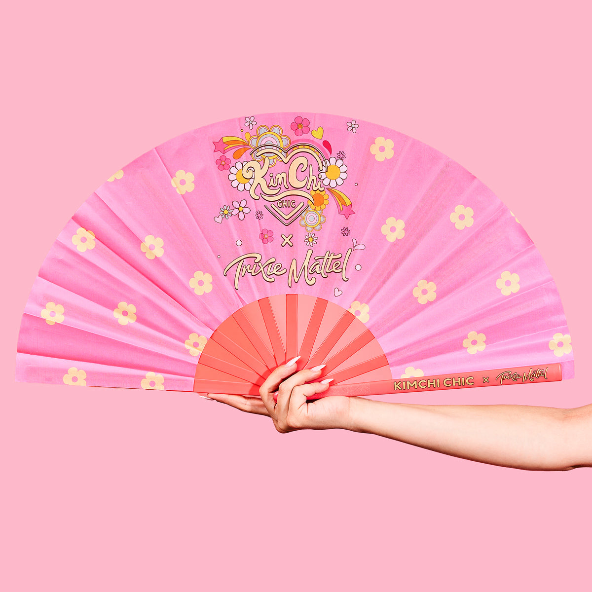 Hand holding a pink decorative fan with floral designs against a pink background.