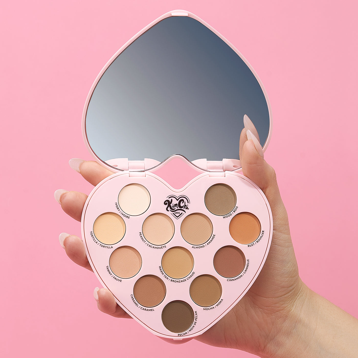 Hand holding heart-shaped makeup palette with mirror on pink background