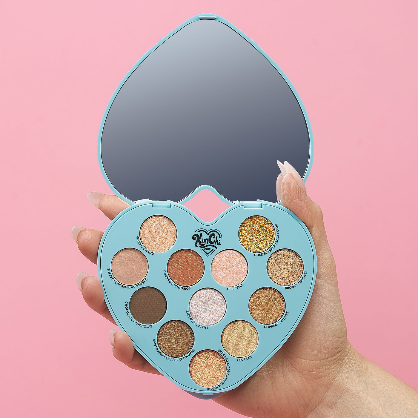 Hand holding a heart-shaped eyeshadow palette with a mirror against a pink background.