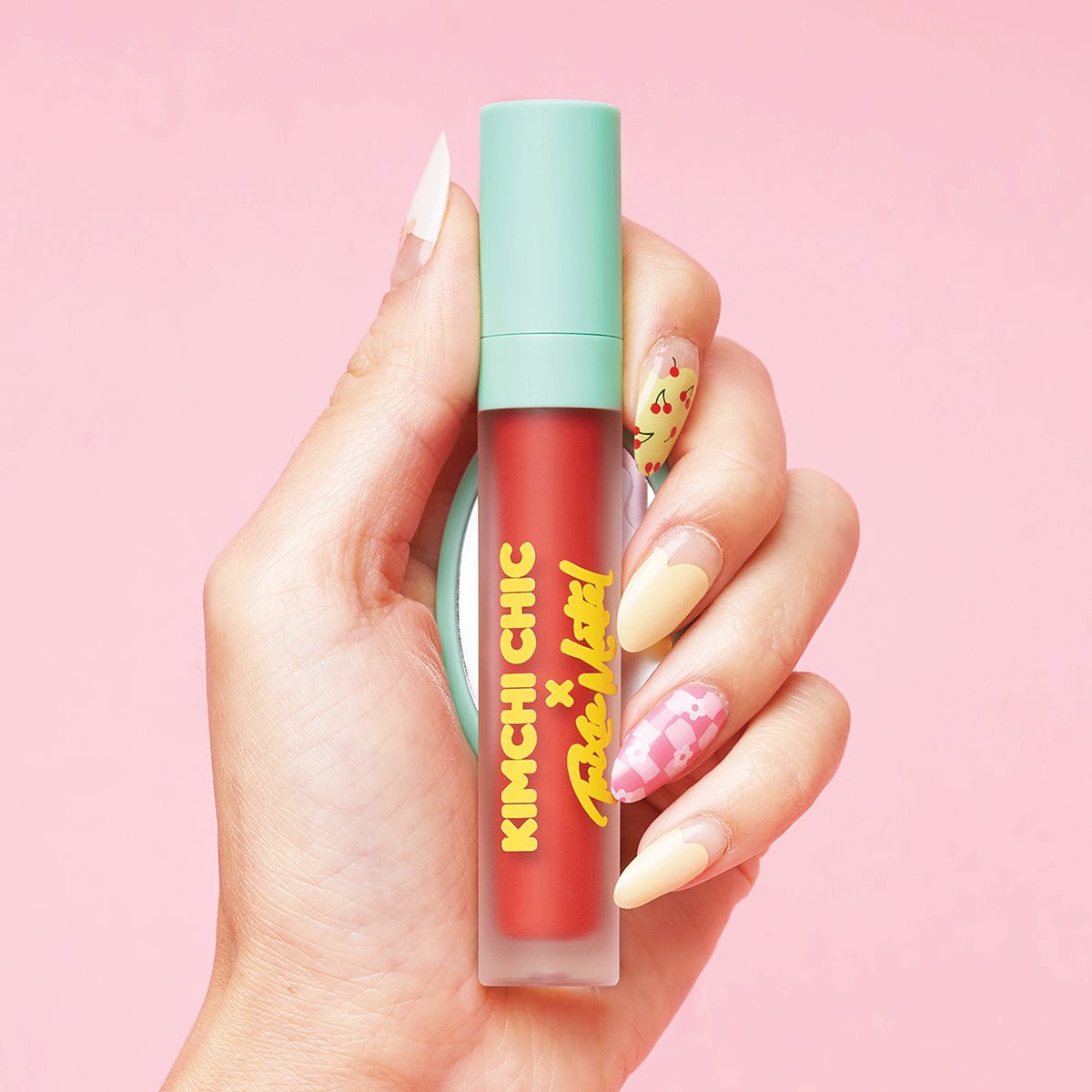 Hand holding a red lipstick bottle with mint green cap on a pink background.