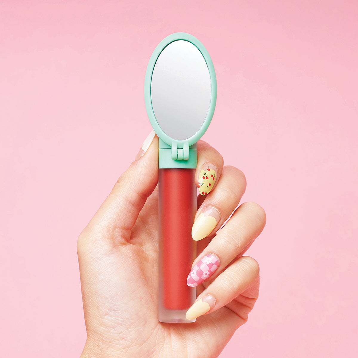 Hand holding a red lip gloss with a small attached mirror, against a pink background.