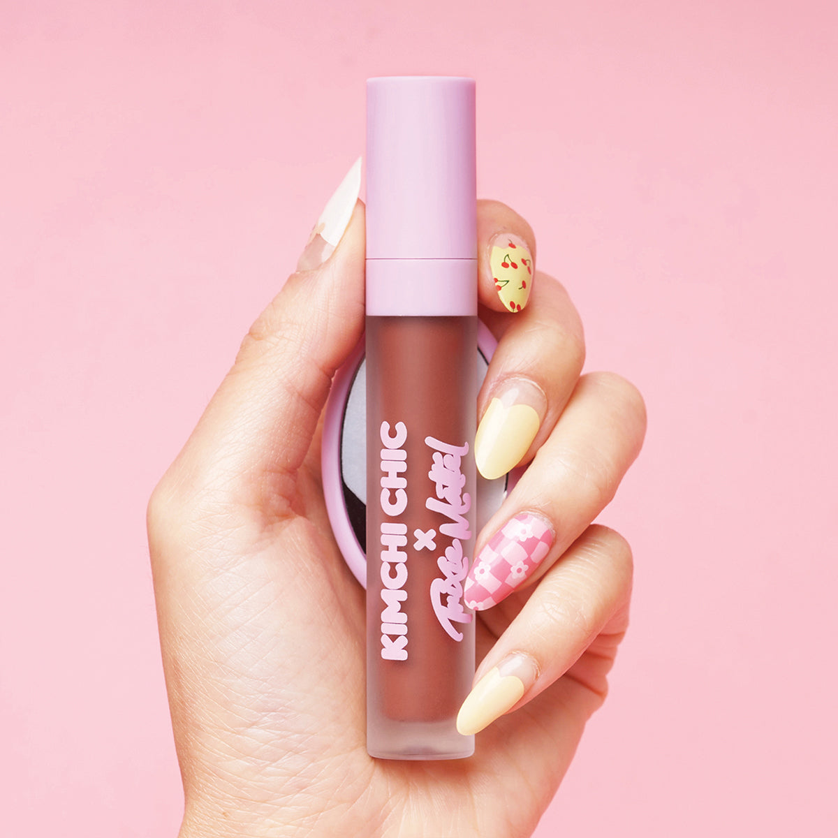 Hand holding a pink-topped lipstick with decorated nails against a pink background.