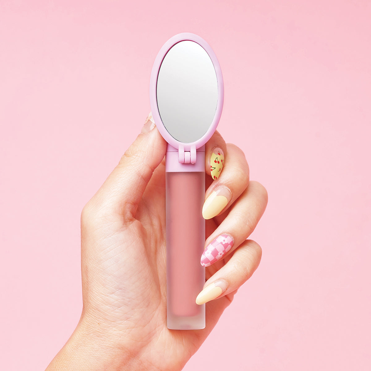 Hand holding a pink lip gloss with a built-in mirror against a pink background.