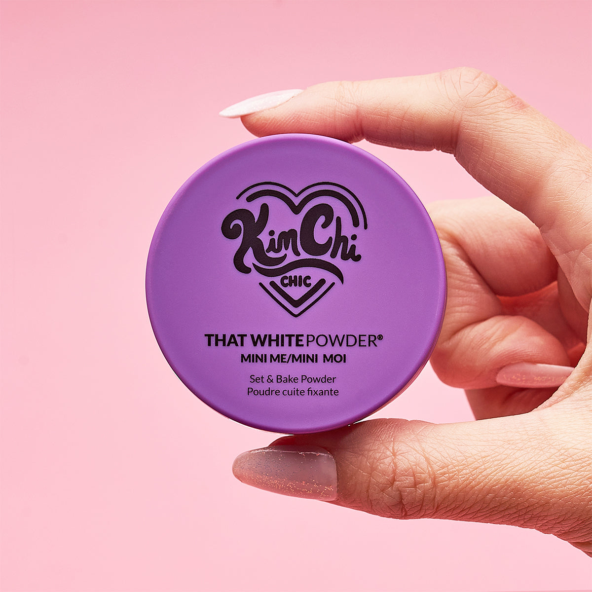 Hand holding a purple container of KimChi Chic beauty powder against a pink background.