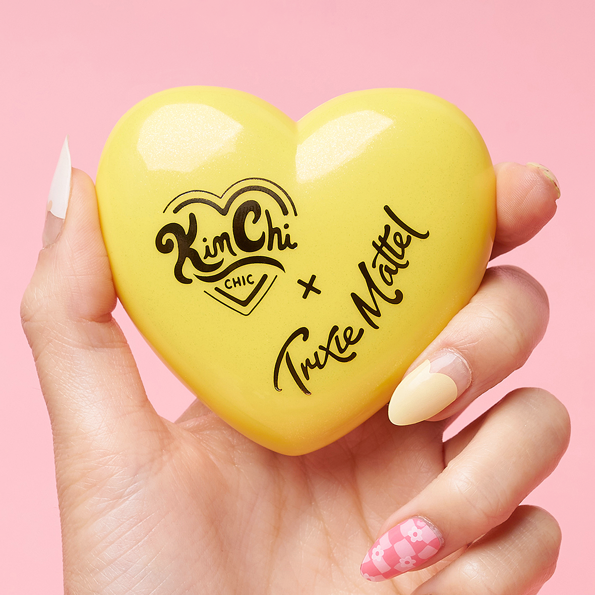 Hand holding a yellow heart-shaped object with 'Kim Chi Chic x Trixie Mattel' written on it.