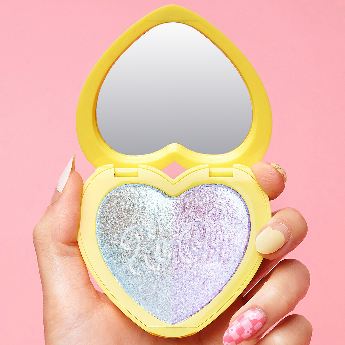 Heart-shaped compact with mirror and iridescent powder held by hand against pink background.